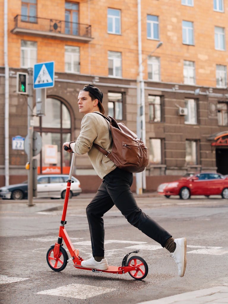 Man in Brown Long Sleeve Shirt with Backpack Riding a Red Kick Scooter